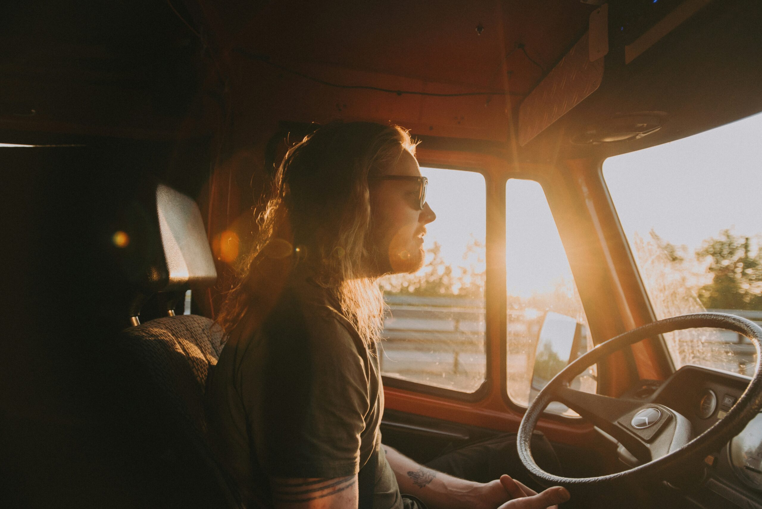 a long-haired truck drivers sits behind the wheel, one thumb lightly touching the bottom of the steering wheel, as the sun shines low in the distance