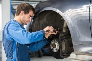 Auto mechanic in a blue work uniform inspecting or repairing a car’s exposed wheel hub and brake assembly inside a service garage.
