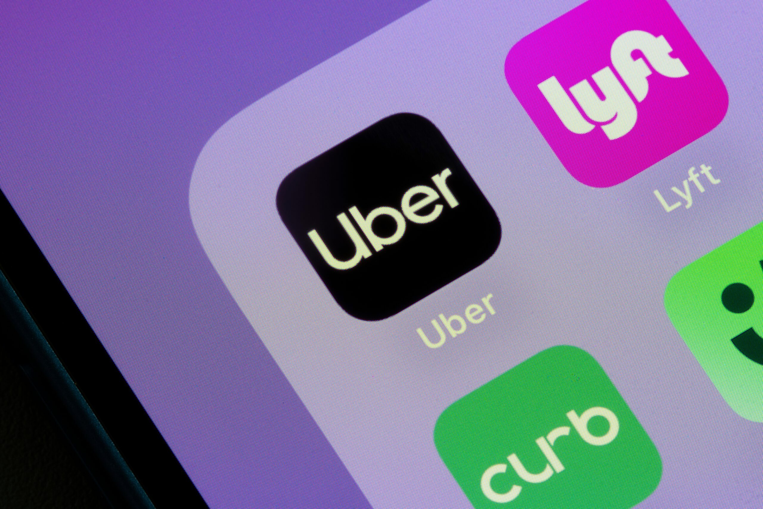 Close-up of a smartphone screen displaying ride-hailing app icons, with the Uber app logo centered in black and white, alongside Lyft and other transportation apps on a softly lit, purple-tinted background.