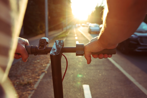 First-person view of hands gripping the handlebars of an electric scooter while riding along a road at sunset, with cars visible ahead and warm sunlight flaring across the scene.