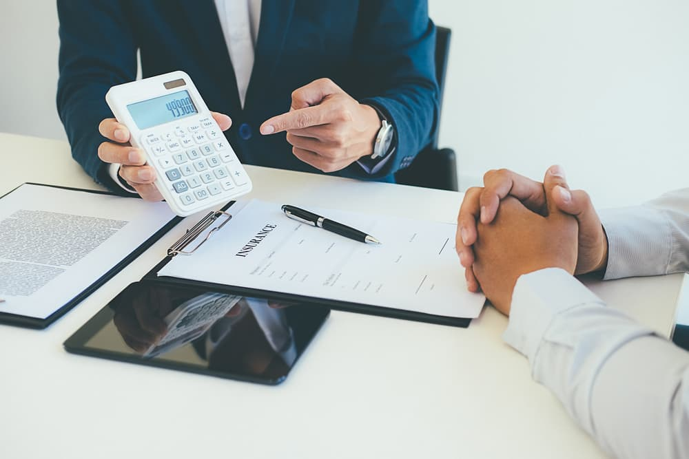 Insurance agent points to a calculator while discussing policy costs across a desk with a client, with insurance documents, a pen, tablet, and clipboard visible.