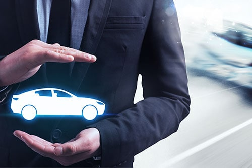 Business professional in a suit holds a glowing car icon between their hands, symbolizing vehicle protection or auto insurance against a blurred roadway background.