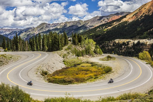 A wide, scenic mountain landscape with a sharply winding paved road forming a dramatic hairpin turn through alpine terrain, surrounded by dense evergreen forests, rolling hills, and rugged rocky mountain peaks under a bright sky filled with scattered white clouds, with two small motorcycles riding along opposite curves of the road and patches of yellow-green vegetation in the center of the bend.