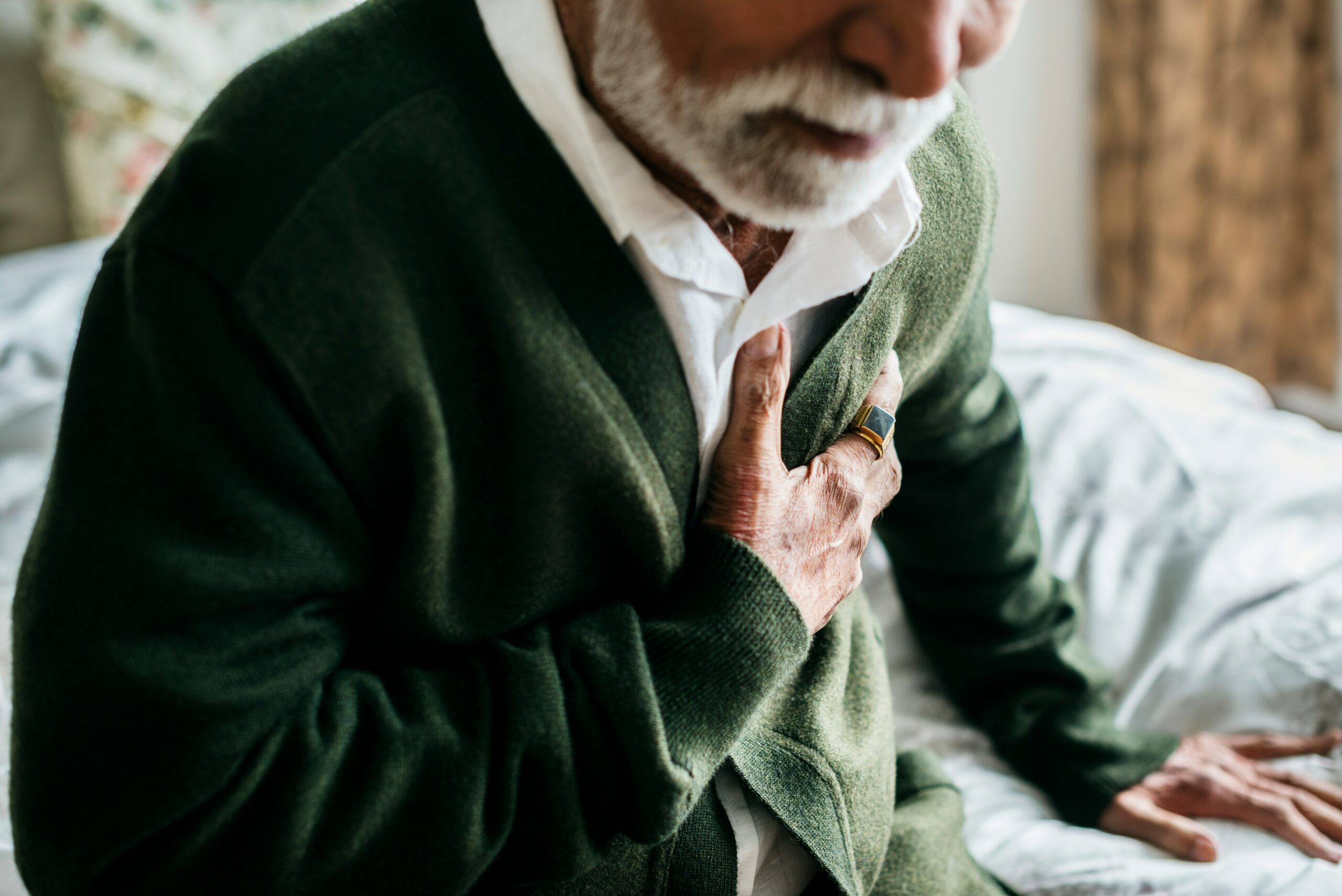 An older man sites on a bed, clutching his chest.