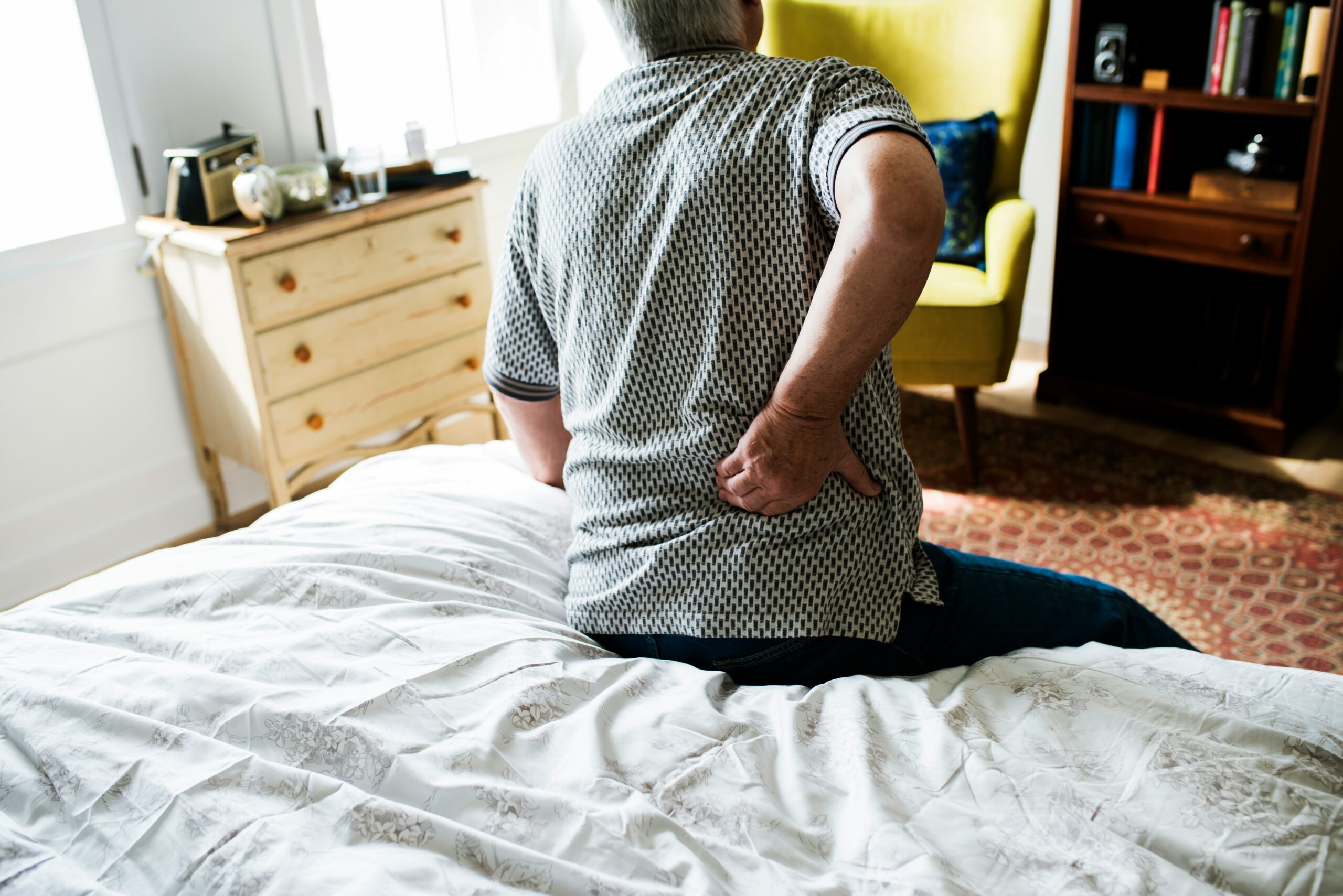 Man sitting on the edge of a bed holding his lower back in pain, suggesting discomfort or a back injury