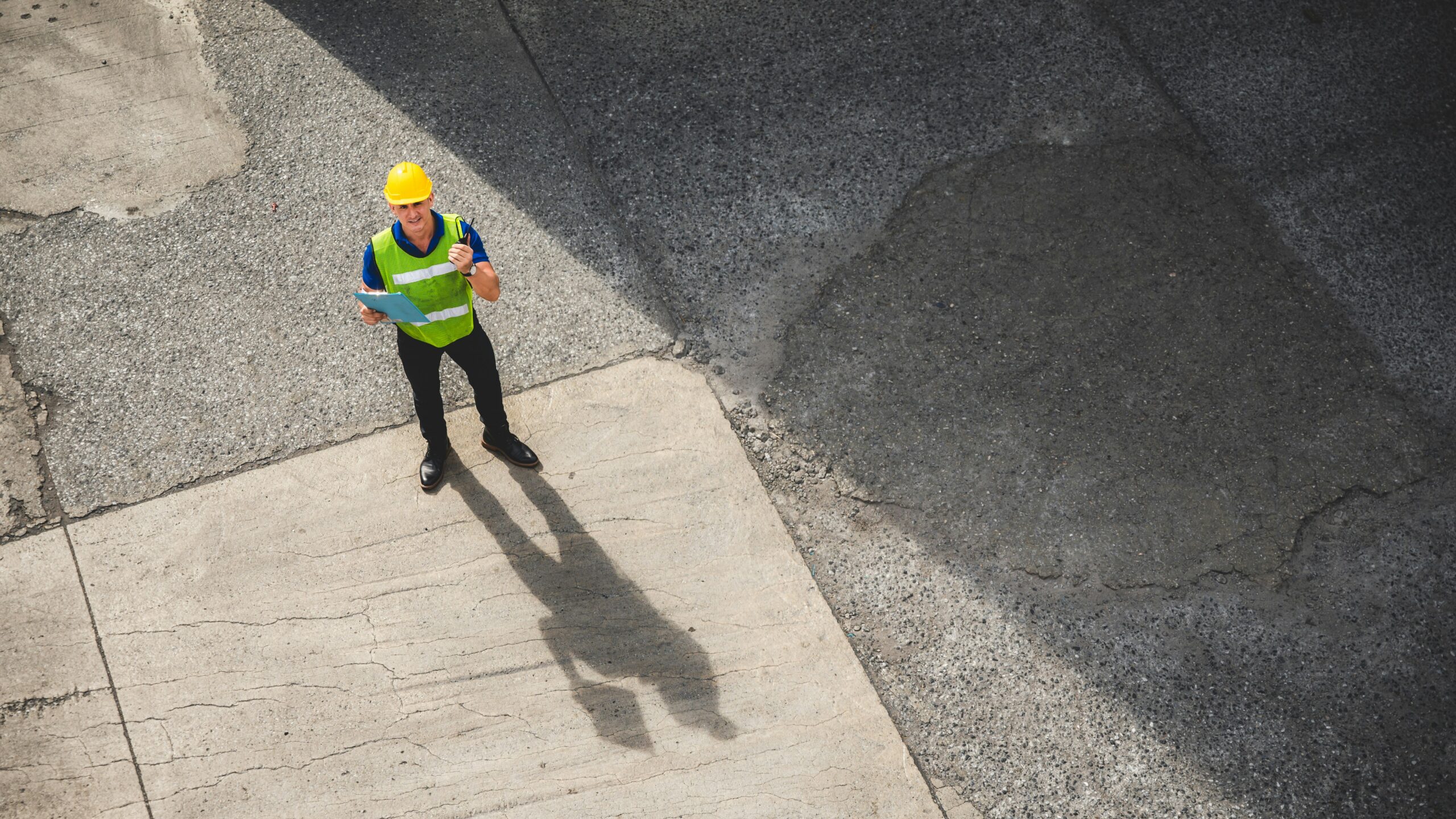 An overhead view of a construction worker standing on a street, wearing a hard hat and safety vest, surrounded by equipment.