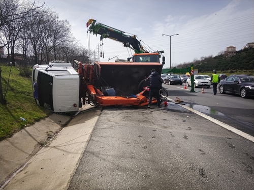 Overturned truck and damaged vehicle on the side of a busy road, with emergency responders and a tow truck working to clear the scene of a traffic accident.