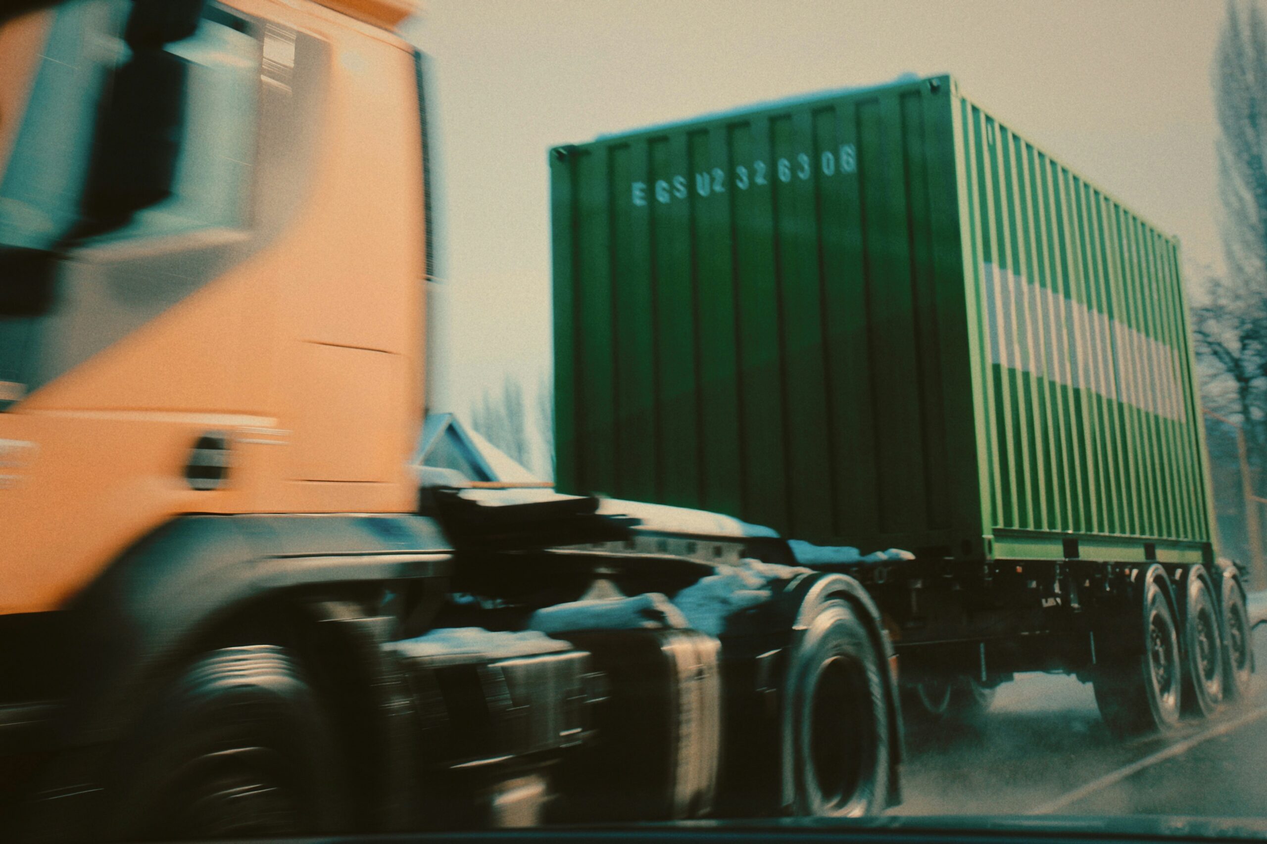 A truck drives on a road, carrying a green container in its cargo area.