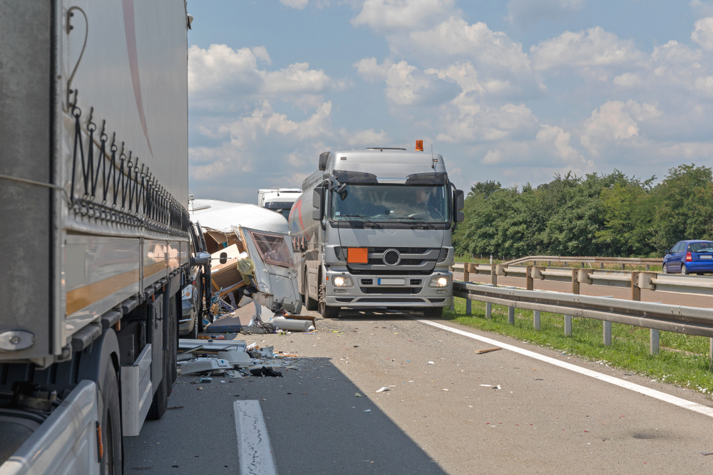 A truck drives along a road, surrounded by trees and open fields under a clear blue sky.