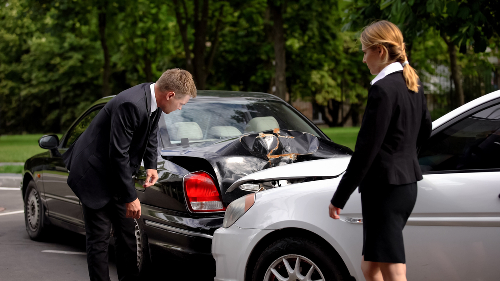 Two people in business attire inspecting vehicle damage after a rear-end car accident in a parking lot with visible damage to both cars