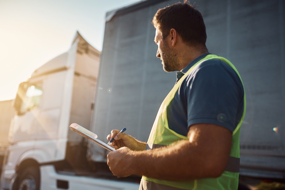 A man in a safety vest is writing notes on a clipboard while standing in an outdoor setting.