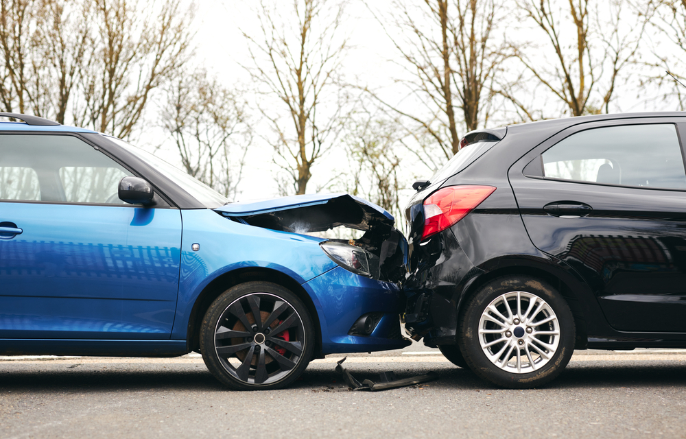 Two cars damaged in a collision, with visible dents and broken parts, parked on the side of the road.