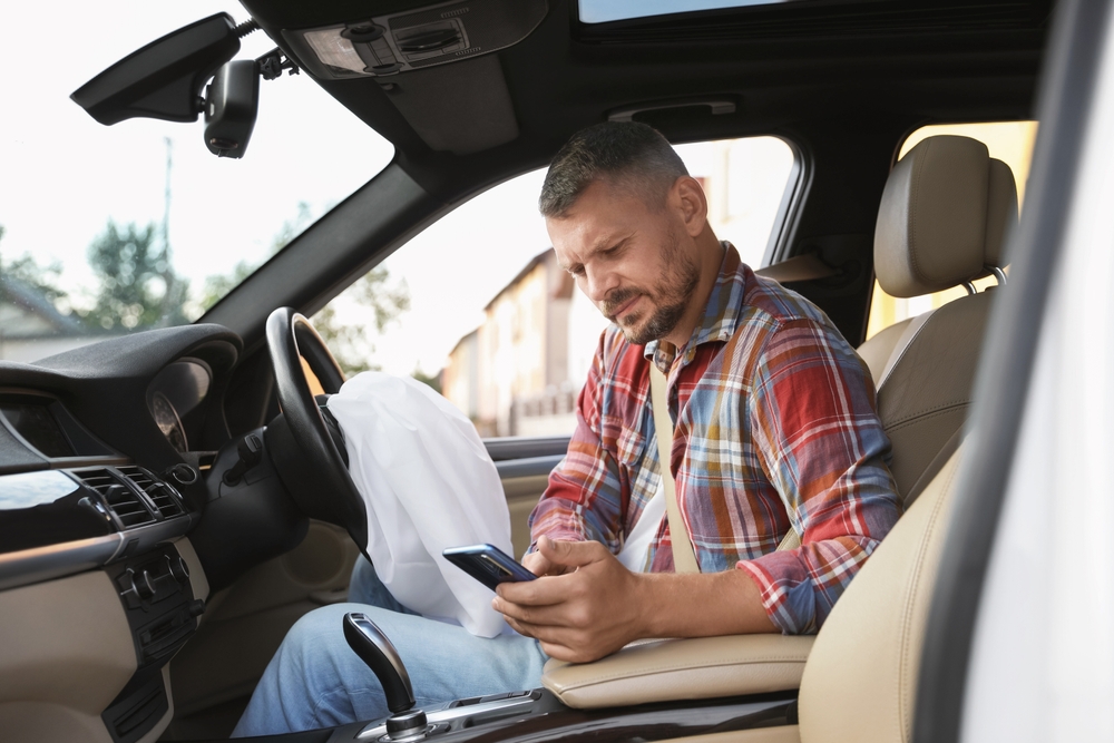 Man checking his phone next to a deployed airbag after a car accident